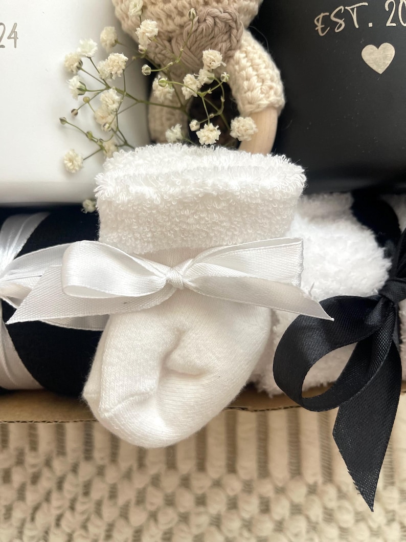 May include: A white baby sock with a white satin bow tied around it. The sock is sitting on top of a black and white striped ribbon. The sock is part of a baby gift basket.
