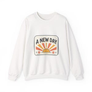 May include: Cream-coloured sweatshirt with a crew neck. The front displays a rectangular graphic with the text "A NEW DAY" above a stylized sunrise in orange and yellow. Small floral accents are on either side of the sunrise.