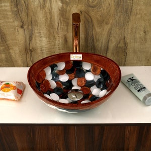 May include: Oval-shaped bathroom sink with a brown, white, and black pebble design. The sink is made of resin and has a copper-colored faucet.