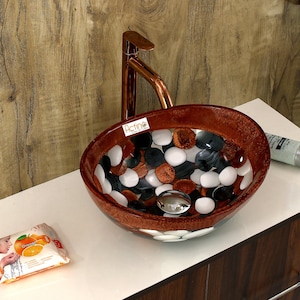 May include: A brown oval-shaped bathroom sink with a white and black pebble design. The sink has a copper faucet and a chrome drain.