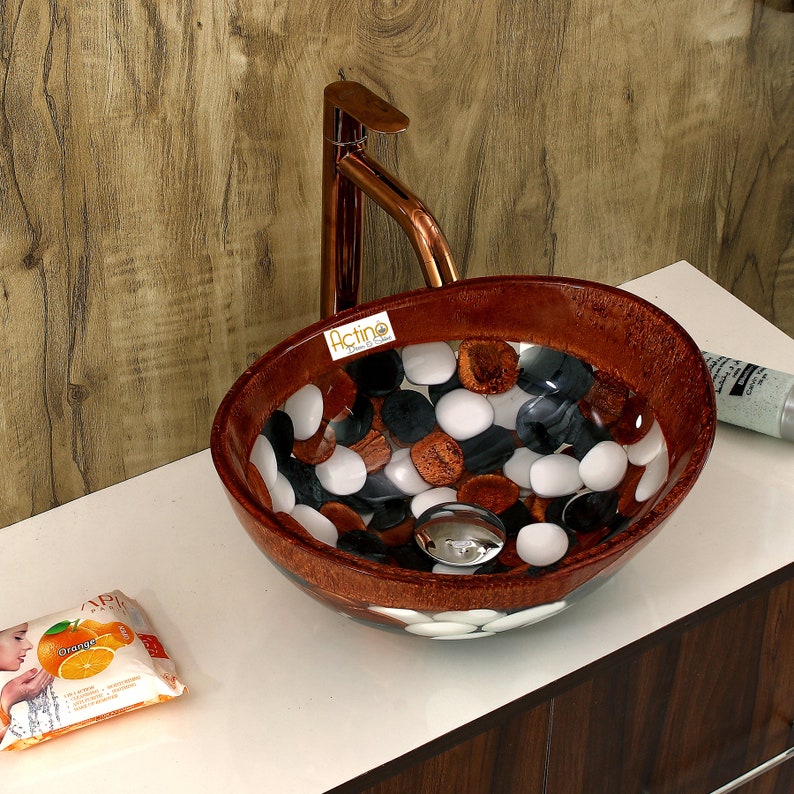 May include: A brown oval-shaped bathroom sink with a white and black pebble design. The sink has a copper faucet and a chrome drain.