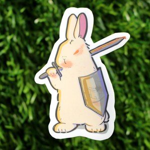May include: A die-cut sticker featuring a cream-colored rabbit standing upright. The rabbit holds a long, thin, multi-colored pencil across its shoulders like a sword and carries a gold-toned shield with the text MOCHAMOCHA printed on the front.