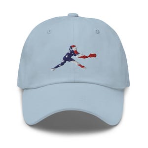 Può includere: Un berretto da baseball blu chiaro con un ricamo a forma di persona che gioca a baseball. La persona è in silhouette nei colori della bandiera americana.