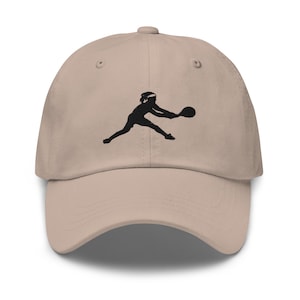 Può includere: Un cappello da baseball beige con una silhouette nera di una persona che gioca a tennis ricamata sul davanti.