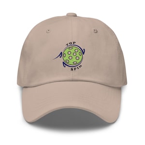 Op de afbeelding: Een beige baseballpet met een geborduurd logo van een groene pickleball met de tekst "TOP SPIN" in blauw.