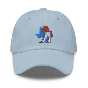 Op de afbeelding: Een lichtblauwe baseballpet met een geborduurd ontwerp van een golfer die een club zwaait voor de omtrek van de staat Texas. De golfer draagt een rood shirt en een witte broek.