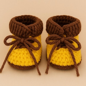 Puede incluir: Un par de patucos de bebé hechos a mano con ganchillo. Los patucos son amarillos con ribetes y cordones marrones. Los cordones están atados con lazos. Los patucos están diseñados para parecerse a zapatos en miniatura.