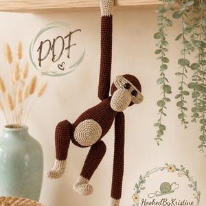 Könnte beinhalten: Ein handgehäkelter Affe in Braun und Beige, der an einem Holzregal hängt. Der Affe hat einen runden Körper, lange Arme und Beine und ein lächelndes Gesicht. Das Bild enthält den Text "PDF" und ein Logo mit der Aufschrift "HookedByKristine".