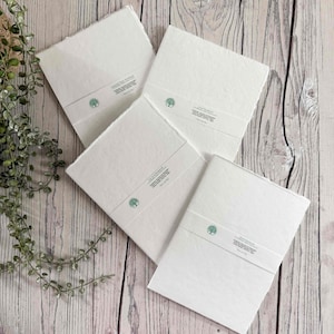 Könnte beinhalten: Vier quadratische Notizbücher mit strukturierten weißen Papierumschlägen und Büttenrändern. Jedes Notizbuch ist mit einer weißen Papierbanderole umwickelt, die ein kleines grünes Baumsymbol und den Text "Seed Paper Notebook" aufweist.