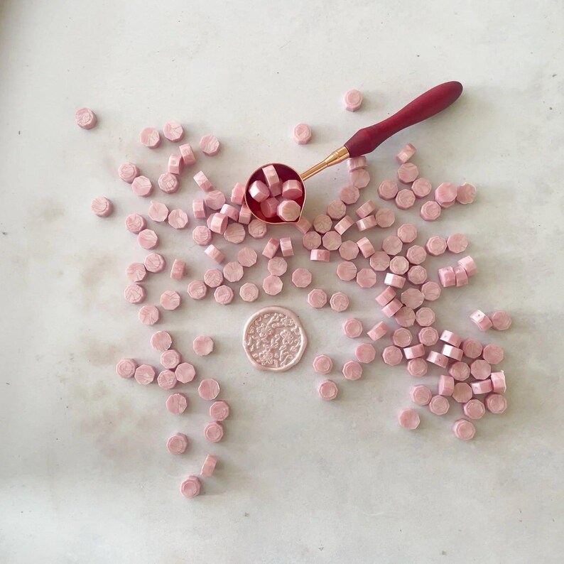 A collection of soft pink, pearlised sealing wax beads scattered on a surface with a red melting spoon above them.