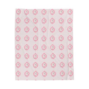 May include: A light pink blanket featuring a repeating pattern of a baby's face with pink hair and a pink bow. The baby's face is drawn in a simple, cartoon style, with a small smile and closed eyes.