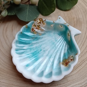 Könnte beinhalten: Muschelförmige Schale mit glänzendem Finish. Die Schale hat eine Mischung aus Weiß und Türkis, mit Goldflocken in dem Harz. Die Ränder sind weiß. Auf einer Holzoberfläche platziert, ideal für Schmuck.