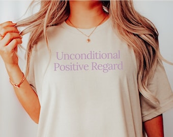 May include: A beige oversized t-shirt with the text "Unconditional Positive Regard" printed in purple on the front.