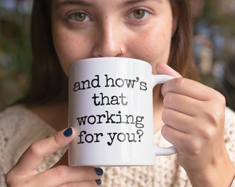 May include: White ceramic mug with the text "and how's that working for you?" in black. The mug is held up to the face, with the handle on the right side. The person is wearing a cream-colored sweater.