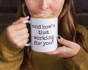 Peut inclure: Une tasse en céramique blanche avec le texte "and how's that working for you?" en noir. La tasse est remplie d'une boisson recouverte de mousse. La personne porte un pull vert.