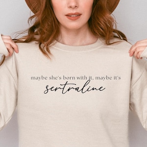 May include: A beige sweatshirt with the text "maybe she's born with it, maybe it's sertraline" printed on the front.