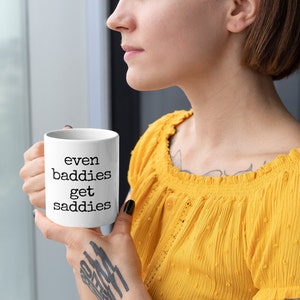 以下が含まれることがあります： 「even baddies get saddies」の文字が黒で書かれた白いセラミックマグ。 黄色のトップスを着た人がマグカップを持っています。 マグカップはコーヒーや紅茶の標準サイズです。