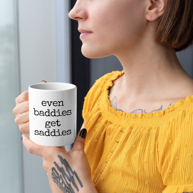 以下が含まれることがあります： 「even baddies get saddies」の文字が黒で書かれた白いセラミックマグ。 黄色のトップスを着た人がマグカップを持っています。 マグカップはコーヒーや紅茶の標準サイズです。