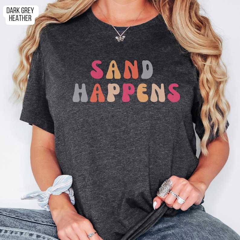 May include: A dark grey heather t-shirt with the text "SAND HAPPENS" in colorful retro-style lettering.