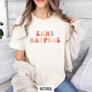 May include: A natural colored t-shirt with the text "SAND HAPPENS" in colorful, retro-style lettering.