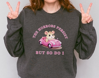 May include: A dark heather grey sweatshirt with a pink cartoon hamster driving a pink car. The text on the sweatshirt reads "The Horrors Persist But So Do I."