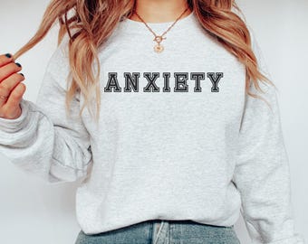 May include: A light gray sweatshirt with the word "ANXIETY" printed in black block letters. The sweatshirt is made of a soft material and has long sleeves. The person is wearing blue jeans.