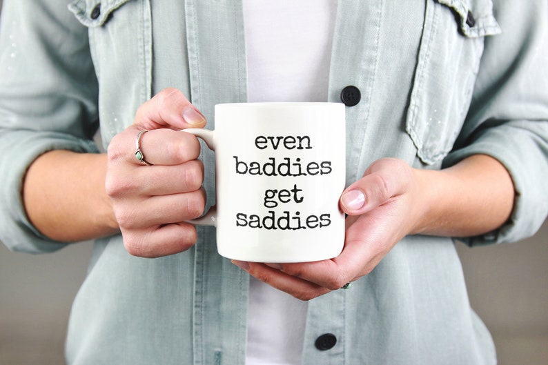 以下が含まれることがあります： 「even baddies get saddies」の黒い文字がプリントされた白いセラミックマグカップ。明るい緑色のジャケットを着た人が持っています。マグカップが中心で、大胆な黒い文字が特徴です。