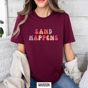 May include: A maroon t-shirt with the text "SAND HAPPENS" in colorful, retro-style lettering.
