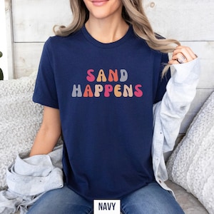 May include: A navy blue t-shirt with the text "SAND HAPPENS" in colorful, retro-style lettering.