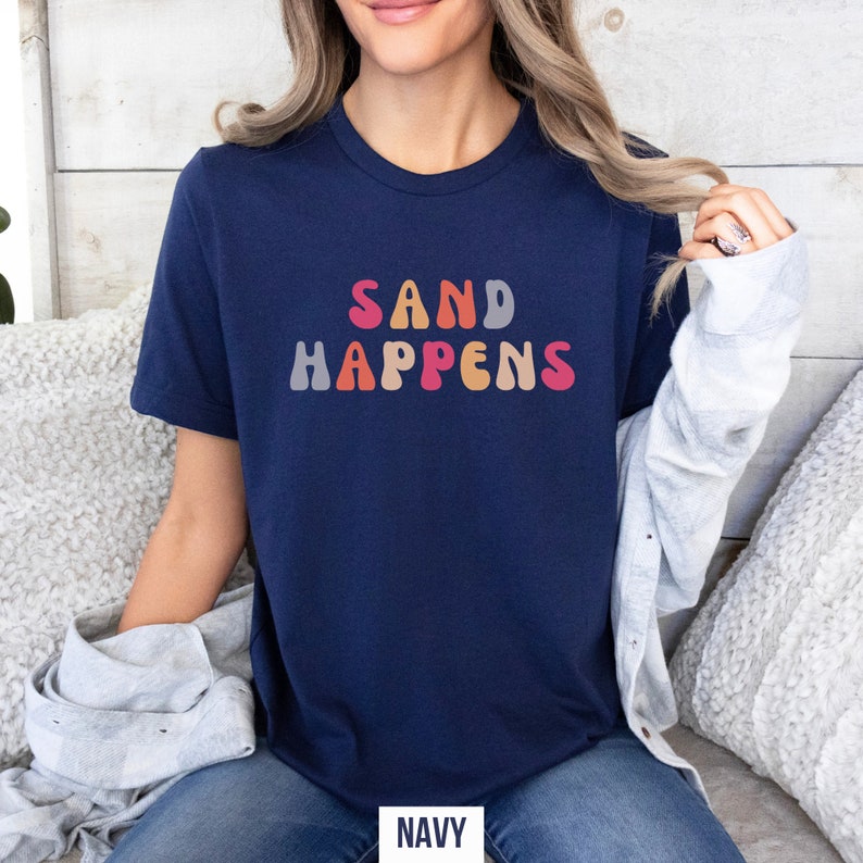 May include: A navy blue t-shirt with the text "SAND HAPPENS" in colorful, retro-style lettering.