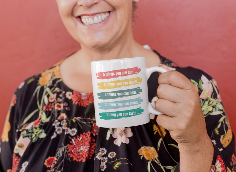 Pu&ograve; includere: Una tazza in ceramica bianca con un design colorato che recita "5 cose che puoi vedere, 4 cose che puoi toccare, 3 cose che puoi sentire, 2 cose che puoi odorare, 1 cosa che puoi assaggiare".
