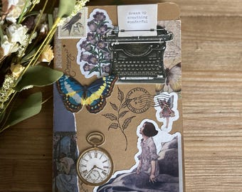May include: A brown notebook with various stickers, including a typewriter, a butterfly, a fairy, a pocket watch, and a quote that reads "dream up something wonderful".
