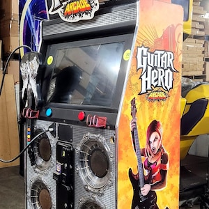 Peut inclure: Une borne d'arcade Guitar Hero avec un grand écran, des haut-parleurs et des accents rouges. L'armoire présente le logo Guitar Hero et un graphisme d'une femme jouant de la guitare. Le jeu est fabriqué par Activision et Konami.
