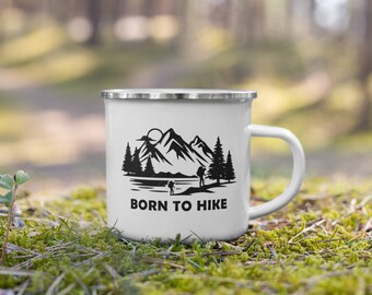 May include: White enamel camping mug with a black silhouette of mountains, trees, and hikers. The mug has a silver rim and handle. The text "Born to Hike" is printed in black below the image.