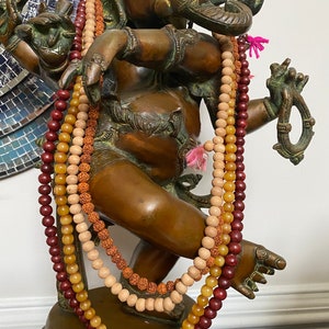 May include: A bronze statue of Ganesha, the Hindu god of wisdom and prosperity, adorned with three beaded necklaces in shades of red, brown, and yellow.