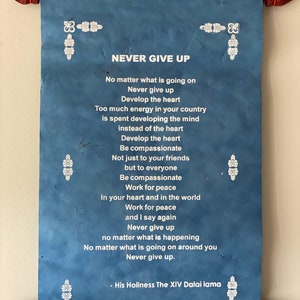 May include: A blue scroll with white text and decorative borders. The text reads "NEVER GIVE UP" and includes a quote from the Dalai Lama: "No matter what is going on, Never give up. Develop the heart. Be compassionate. Work for peace. In your heart and in the world, Work for peace. And I say again, Never give up. No matter what is happening, No matter what is going on around you, Never give up." - His Holiness The XIV Dalai Lama