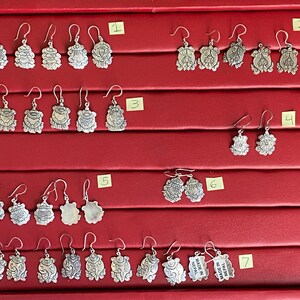 May include: A display of silver earrings with various designs, including floral, geometric, and animal motifs. The earrings are arranged in rows on a red velvet background, with numbers indicating different styles. The text "MADE IN NEPAL" is visible on one pair of earrings.