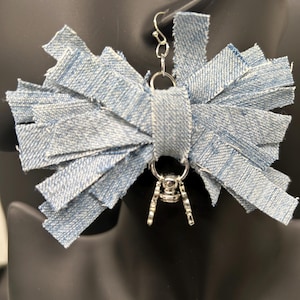 May include: A pair of denim earrings. Each earring features a bow-like design made of strips of light blue denim fabric. The earrings have silver-colored metal hardware, including a hook for the ear and a decorative charm.
