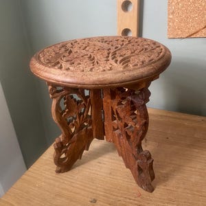 May include: A small, round, brown wooden stool featuring intricate floral carvings on the top surface and openwork, decorative carved legs. The structure is collapsible, with visible metal hinges connecting the two interlocking leg panels.