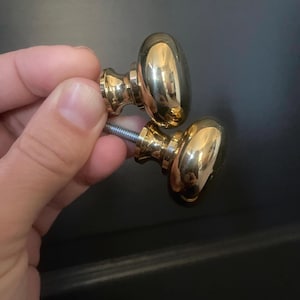 May include: Two polished brass cabinet knobs with an oval shape. The knobs have a shiny, reflective surface and are held by a hand. The knobs are attached to a threaded screw.