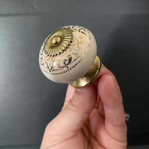 May include: A round ceramic cabinet knob featuring a cream-colored surface with an intricate gold filigree pattern. The center of the knob is adorned with a circular brass-toned metal plate with a sunburst design, and a brass-toned base.