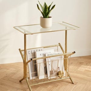 May include: A gold-toned metal side table with a rectangular glass top and a lower wire rack for magazines. A small green snake plant in a cream-colored ceramic pot sits on the glass surface. The rack holds three magazines titled Cereal, Kinfolk, and Architectural Digest.