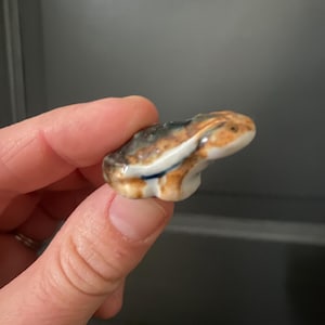 May include: A small ceramic frog figurine. The frog is primarily brown, with white and blue accents. The figurine is held in a person's hand, showcasing its size and detail. The frog's design is detailed, with a smooth, glossy finish.