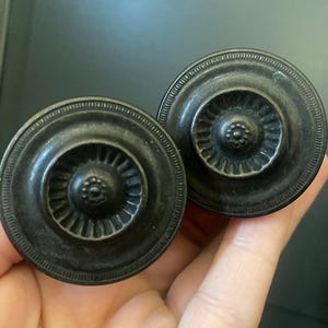 May include: Two antique-style, dark bronze cabinet knobs. Each knob features a circular design with a central floral pattern and a detailed outer rim. These decorative knobs are ideal for adding a vintage touch to furniture.