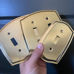 May include: Three polished brass light switch plates with a curved, shield-like shape. Each plate features a central rectangular cutout for a toggle switch and two circular screw holes for mounting, finished with a thin black border.