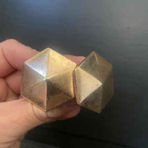 May include: Two gold-colored, geometric cabinet knobs. Each knob has a faceted, hexagonal shape with a slightly worn finish. The knobs are held in a hand, suggesting they are ready for installation on a drawer or cabinet.
