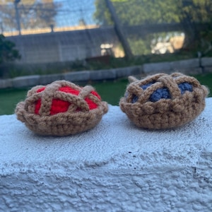 May include: Two crocheted miniature pies. One pie has a red filling, the other has a blue filling. Both pies have a tan crust with a lattice design. The pies are sitting on a white surface.