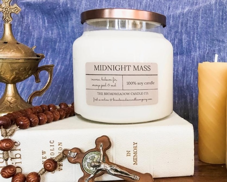 May include: A white glass jar candle with a copper lid. The candle is labeled "Midnight Mass" and has a scent description of "incense, balsam fir, orange peel & musk". The candle is 100% soy and is made by The Broadmeadow Candle Co. The candle is sitting on a white book with the words "In Memory" printed on the spine. There is a wooden rosary and a gold metal incense burner in the background.