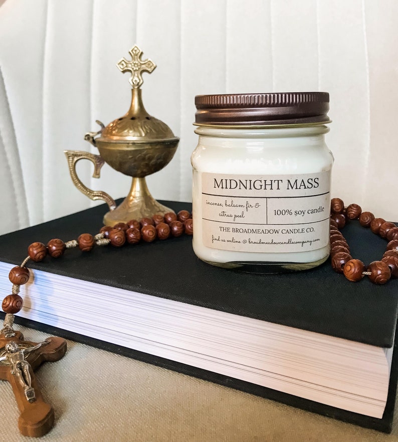 May include: A white soy candle with a brown lid and a label that reads "Midnight Mass" with the scent description "incense, balsam fir & citrus peel" and the company name "The Broadmeadow Candle Co.". The candle is sitting on a black book with a brown rosary and a gold metal incense burner.