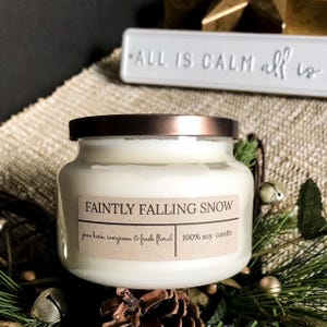 May include: A white soy candle in a glass jar with a copper-colored lid. The label reads "FAINTLY FALLING SNOW" with scent notes of pine bark, evergreen, and fresh floral. The candle is surrounded by pine needles and small bells.